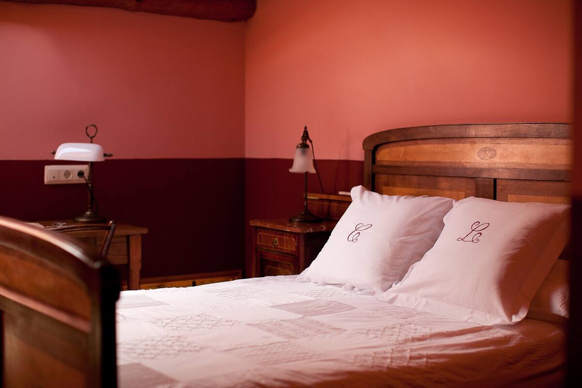 Una cama bien hecha con cabecera de madera, dos almohadas con iniciales bordadas, una mesita de noche con lámpara y una pared con decoración en tonos rojo y blanco.