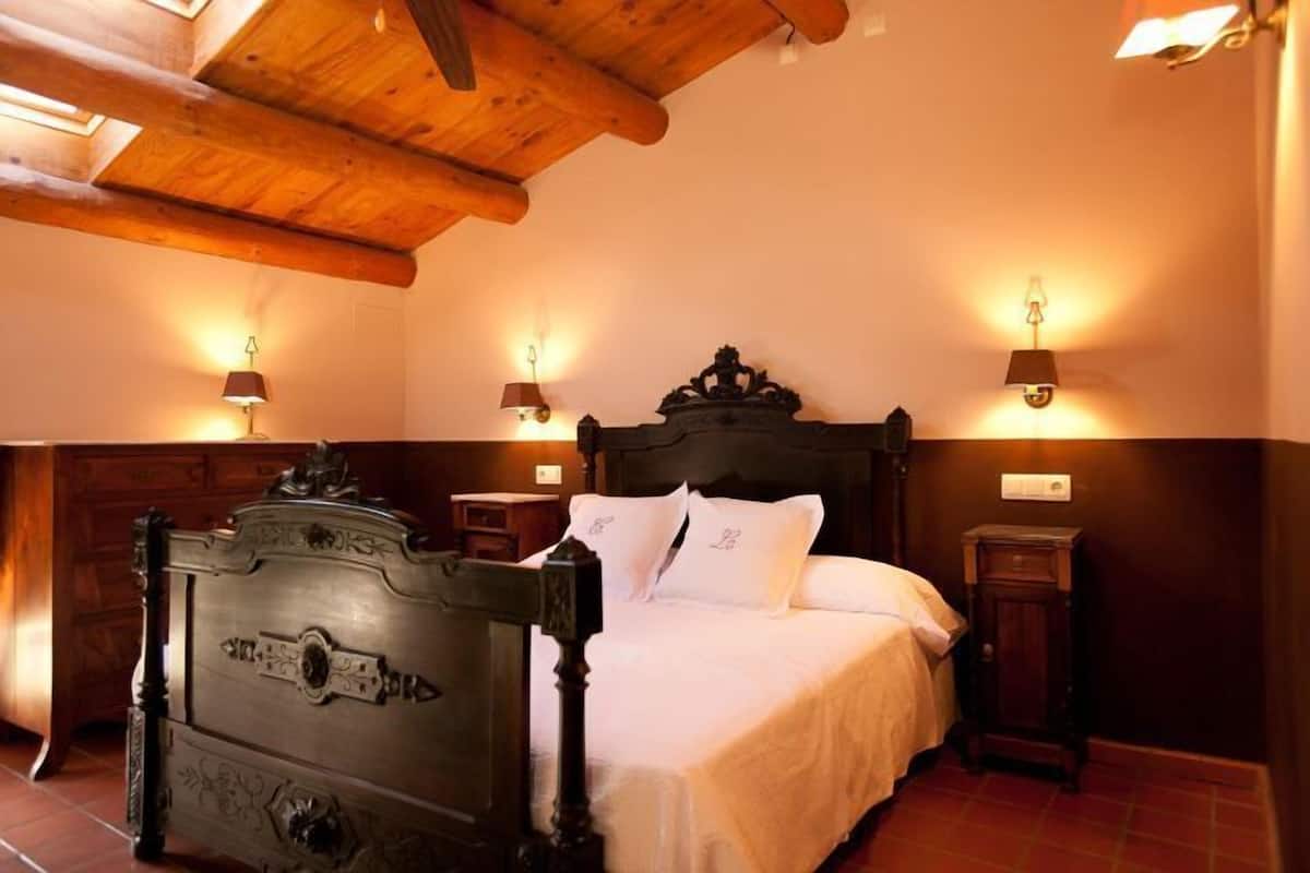 Una cama de madera tradicional con detalles tallados, una mesita de noche y lámparas fijadas a la pared.