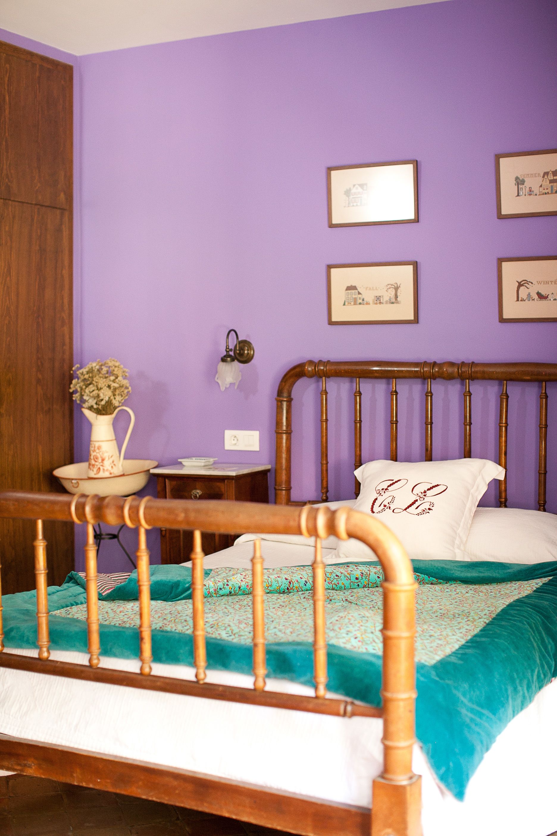 A bedroom with a wooden bed frame, a teal and white bedspread, a white pillow with embroidered text, and a vase with flowers on a nightstand.