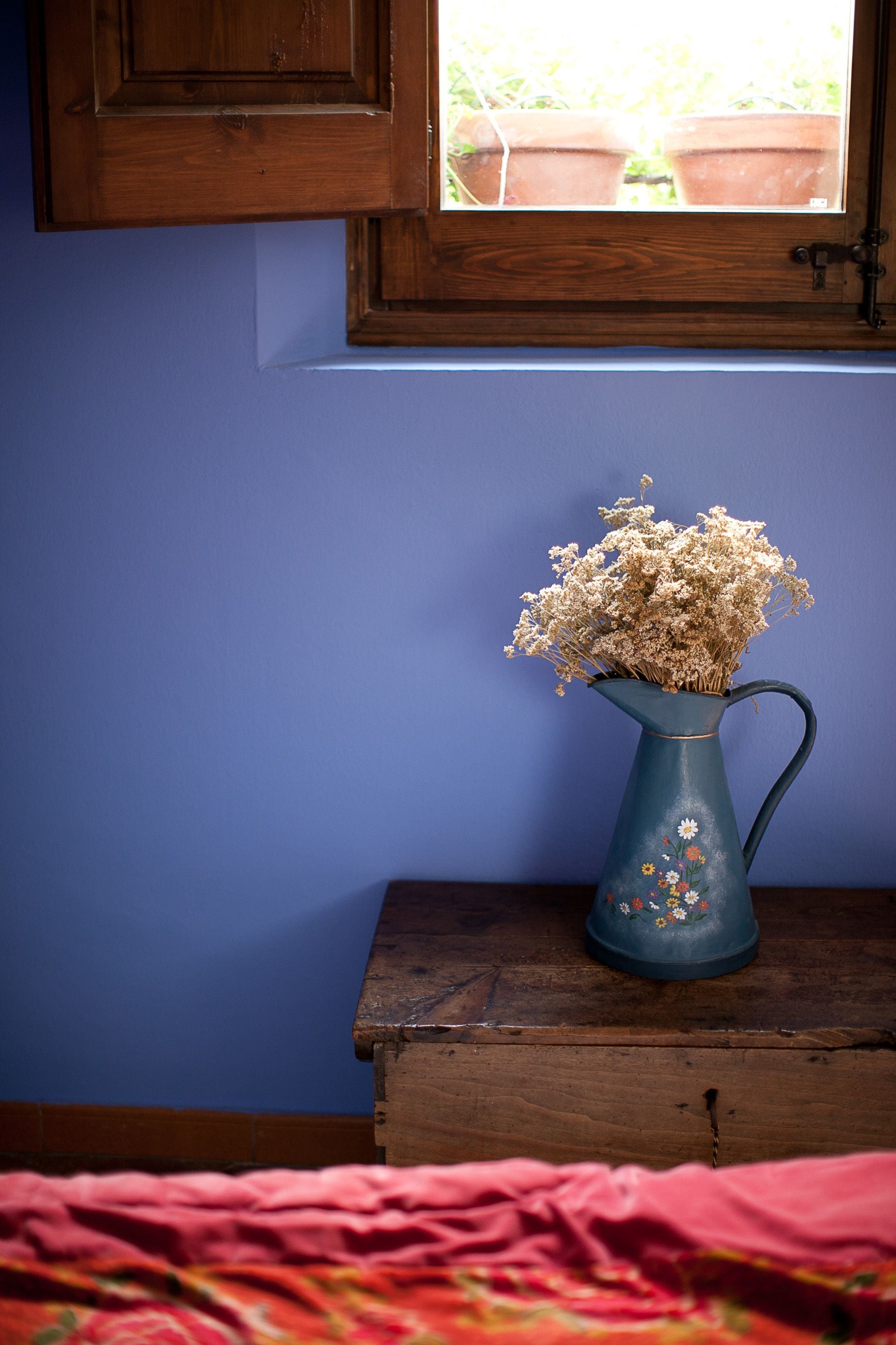 Un jarrón azul con flores secas sobre una mesa de madera cerca de una ventana.