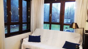 A room with a white sofa, two blue cushions, a wooden table, and large windows with curtains. - El Casar del Puente (Boca de Huergano)