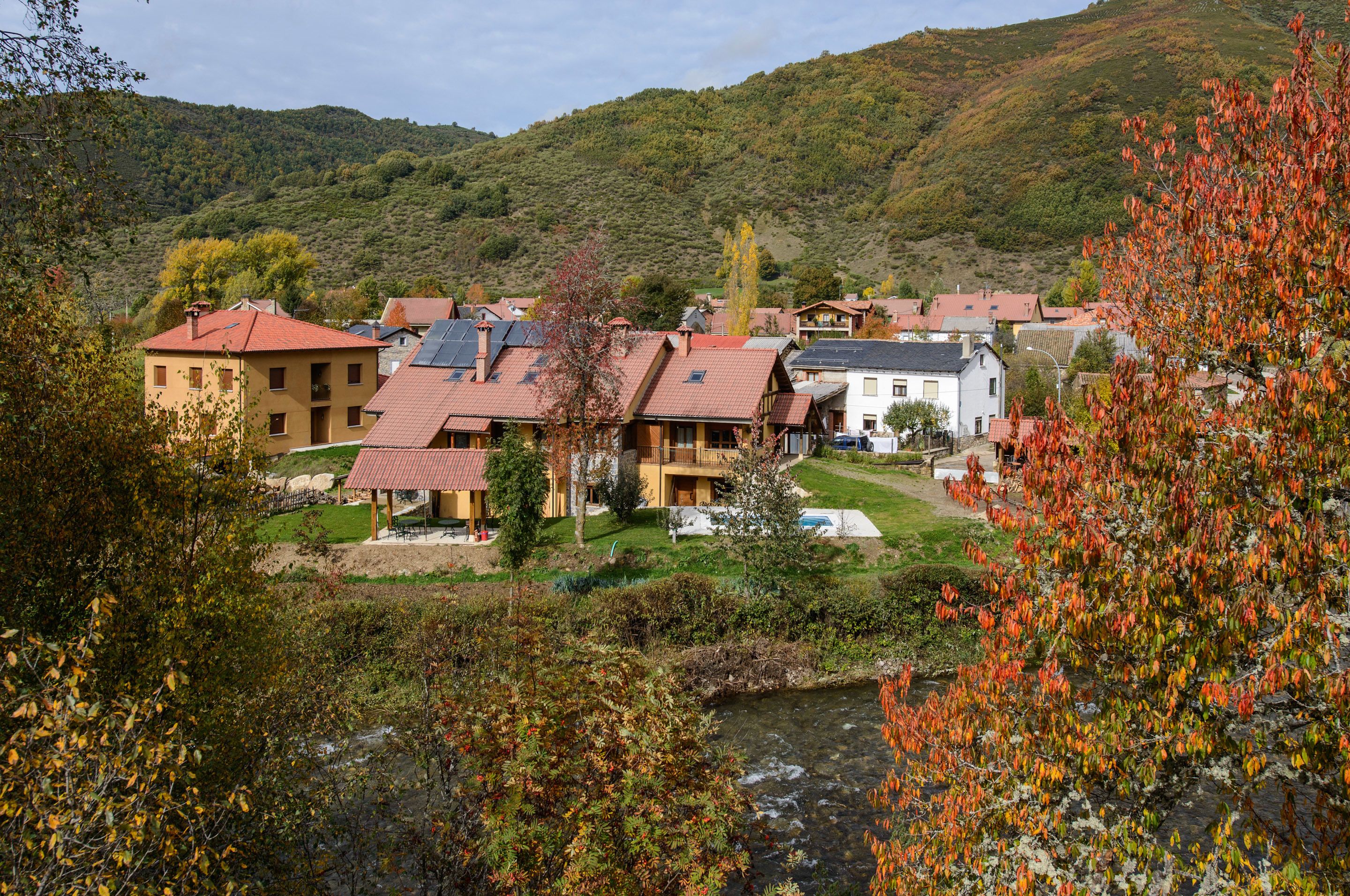 A hillside village with traditional architecture, a river in the foreground, and a mountainous backdrop.