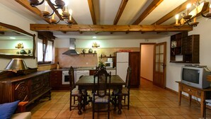 A rustic kitchen with wooden beams, a dining table with chairs, a refrigerator, and a television on a stand. - El Casar del Puente (Boca de Huergano)