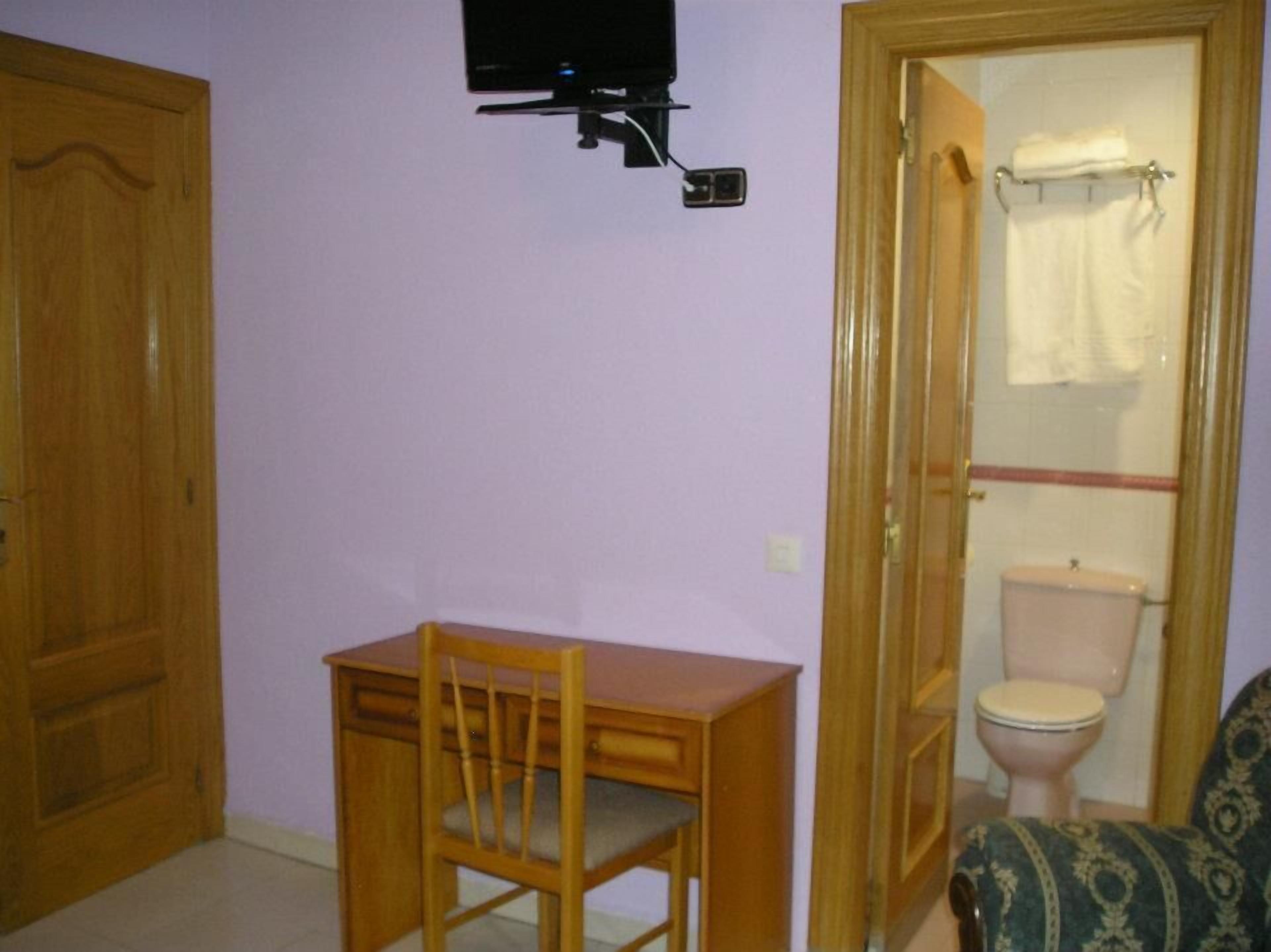 Una habitación con un escritorio de madera, una silla, un televisor montado en la pared y un inodoro a la vista a través de una puerta abierta.