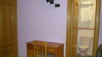 Una habitación con un escritorio de madera, una silla, un televisor montado en la pared y un inodoro a la vista a través de una puerta abierta.