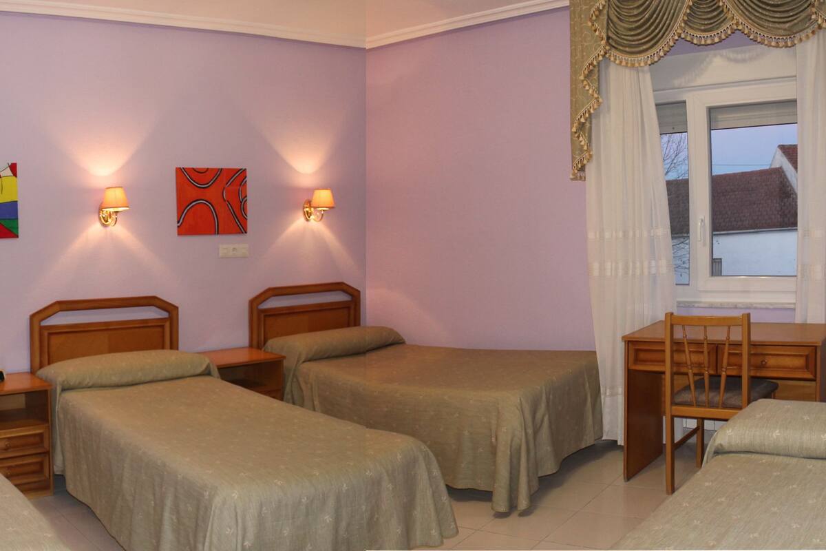 A room with four single beds, a wooden desk, a chair, and two wall-mounted lamps.