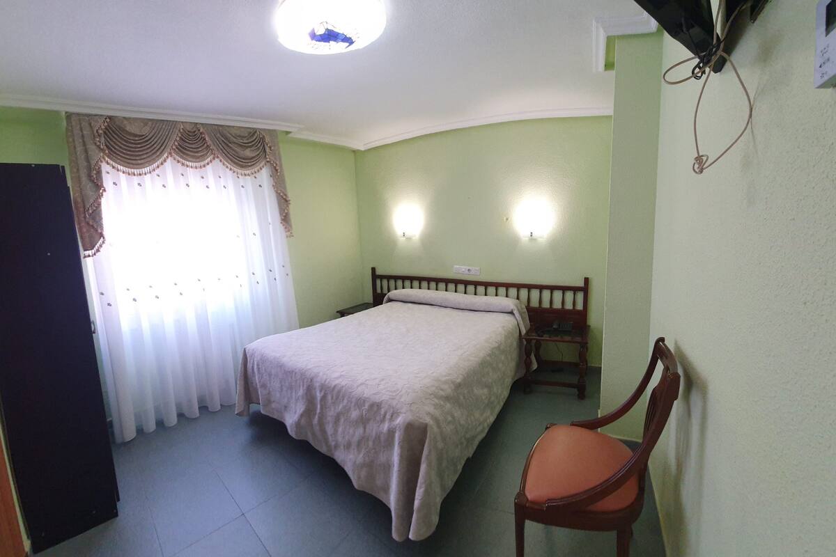 A bedroom with a bed, a chair, a nightstand, a wall-mounted light, and a door.