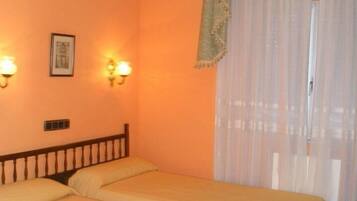 A room with two single beds, a wall-mounted light fixture, a framed picture, and a window with sheer curtains.