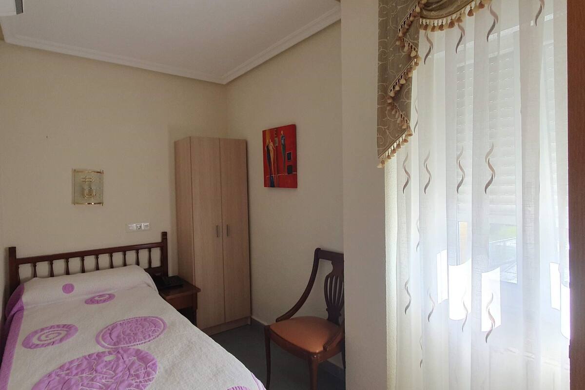 A bedroom with a bed, a wooden wardrobe, a chair, and a door with curtains.