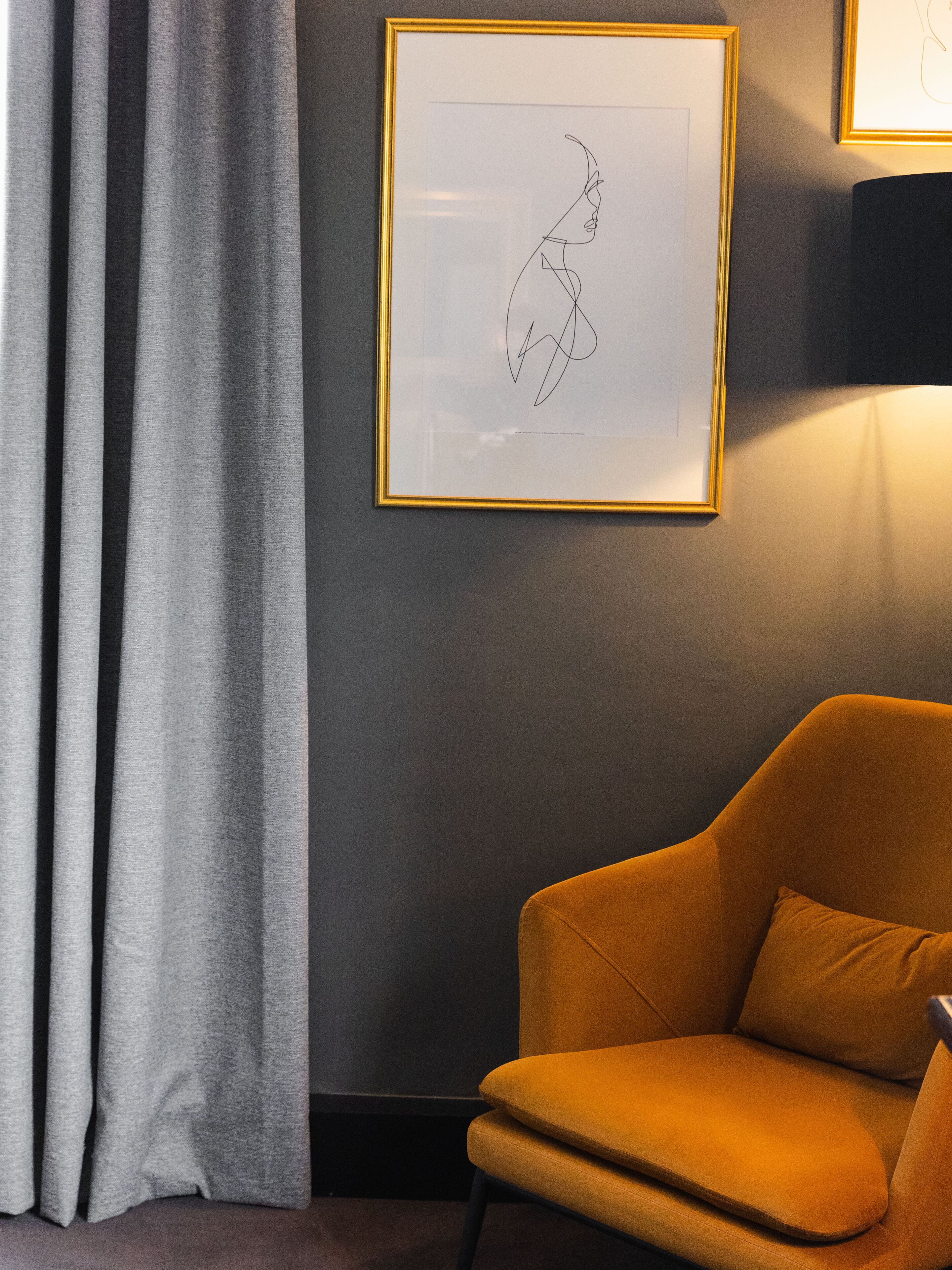 Una habitación con un cuadro abstracto enmarcado en la pared, una cortina gris y un sillón naranja.
