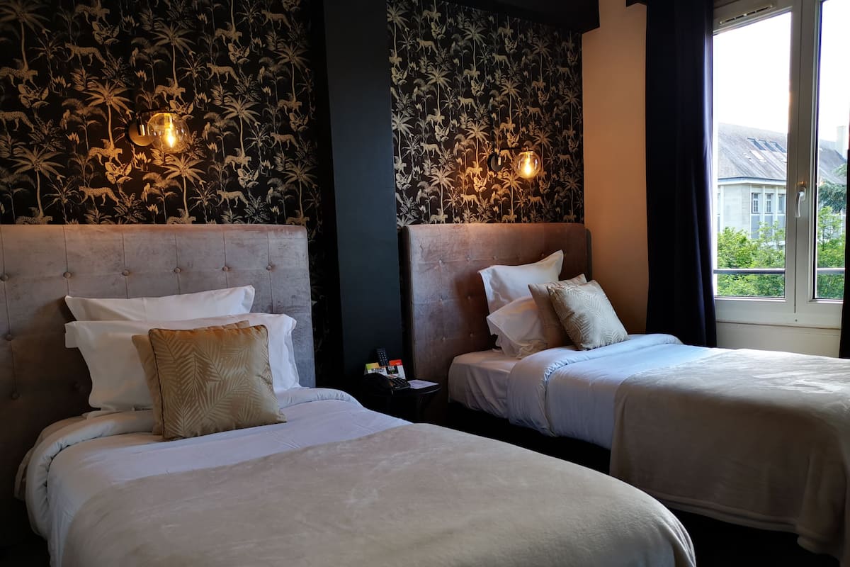 Une chambre d’hôtel avec deux lits, un mur sombre orné d’un motif floral et une fenêtre donnant sur des bâtiments et de la verdure.