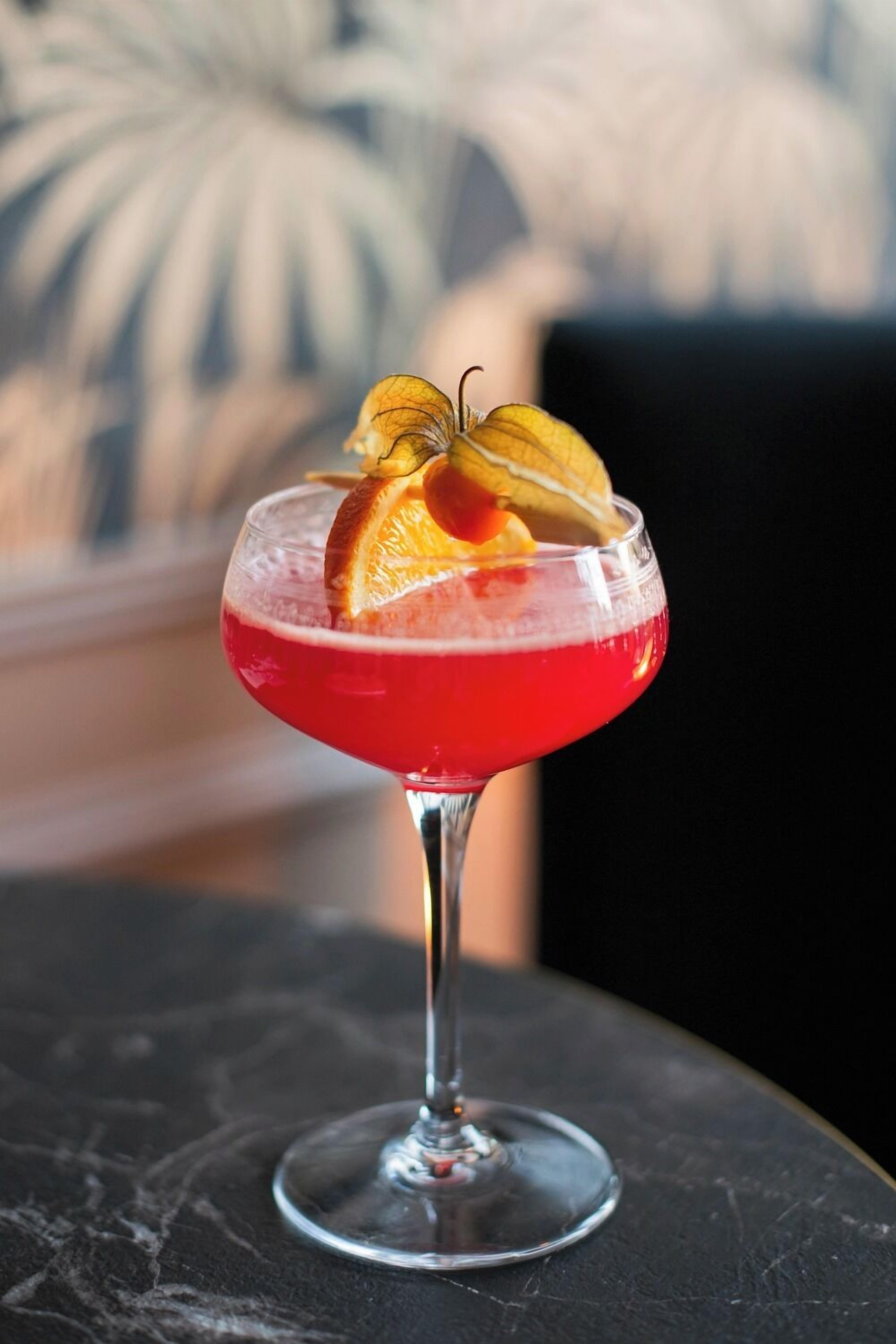 Un cóctel en una copa de tallo con líquido rojo, adornado con una rodaja de naranja y una flor.