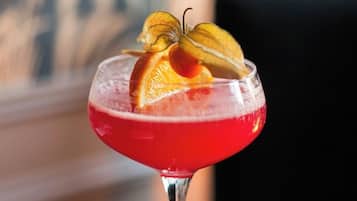 Un cóctel en una copa de tallo con líquido rojo, adornado con una rodaja de naranja y una flor.