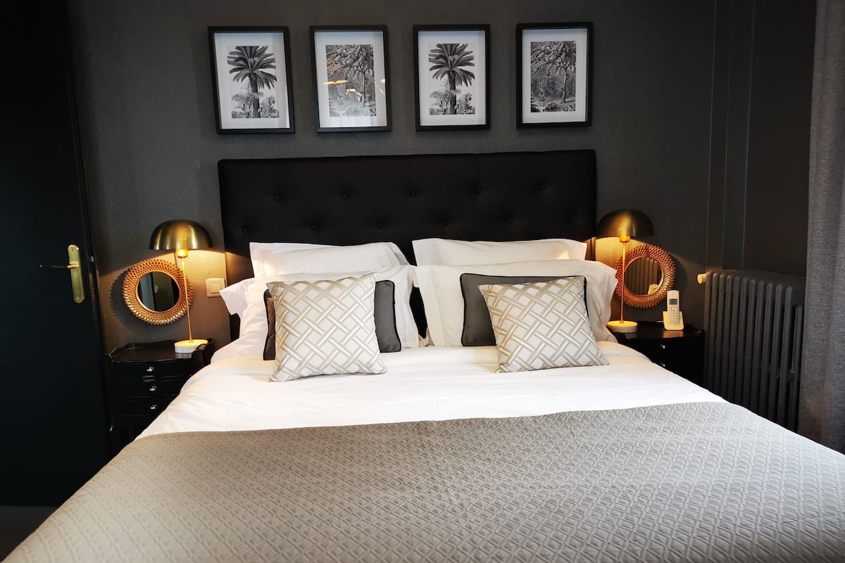 Un lit bien fait, avec du linge de lit blanc et des coussins à motifs, flanqué de deux tables de chevet sur lesquelles sont posées des lampes, et un téléphone fixé au mur.
