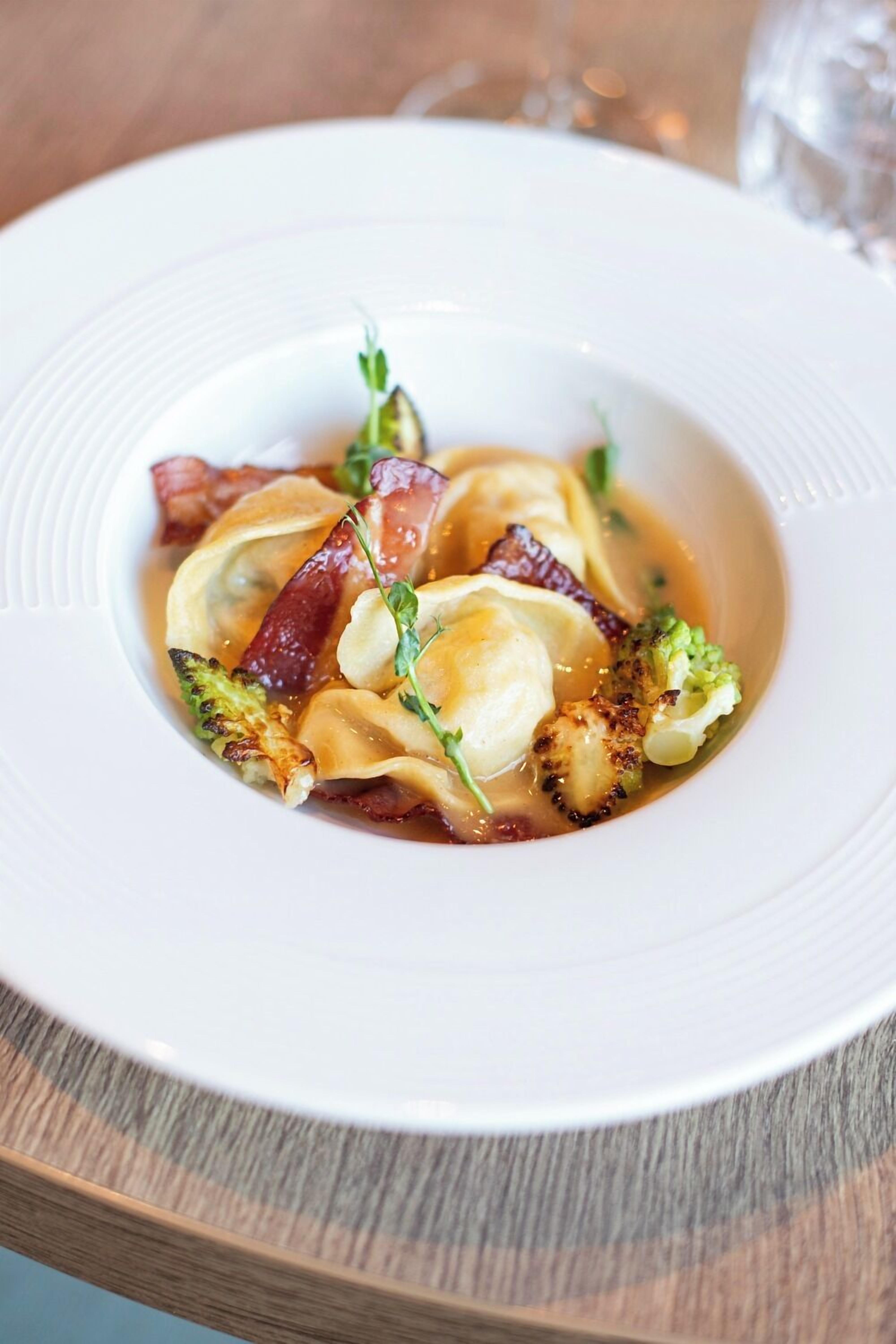 A white plate with ravioli, bacon, and broccoli on a wooden table.