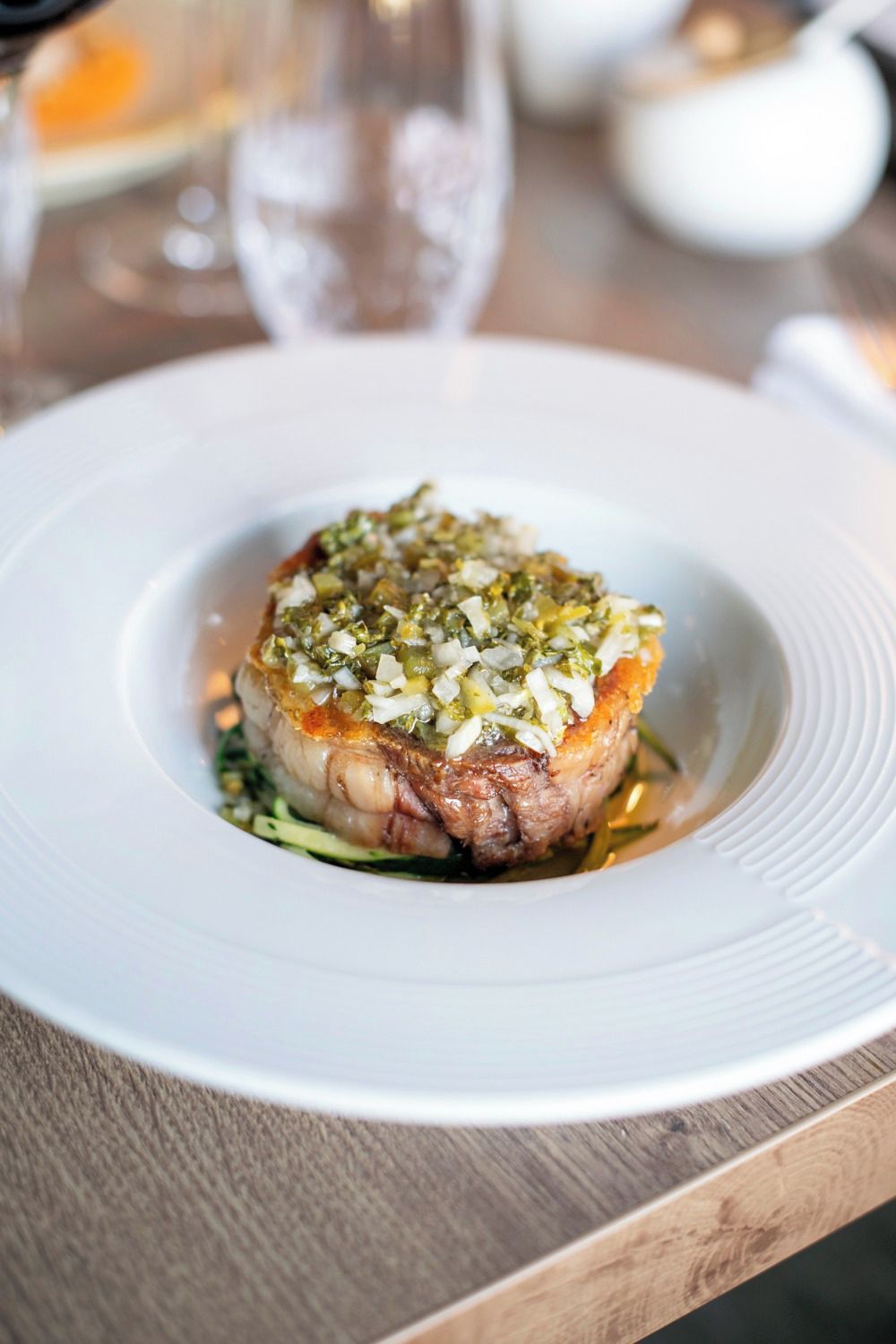 A white plate with a piece of meat topped with a green herb mixture, placed on a wooden table.