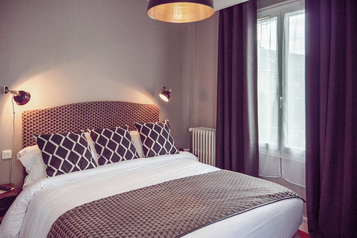 A neatly made bed with patterned bedding, two bedside lamps, and a window with curtains.