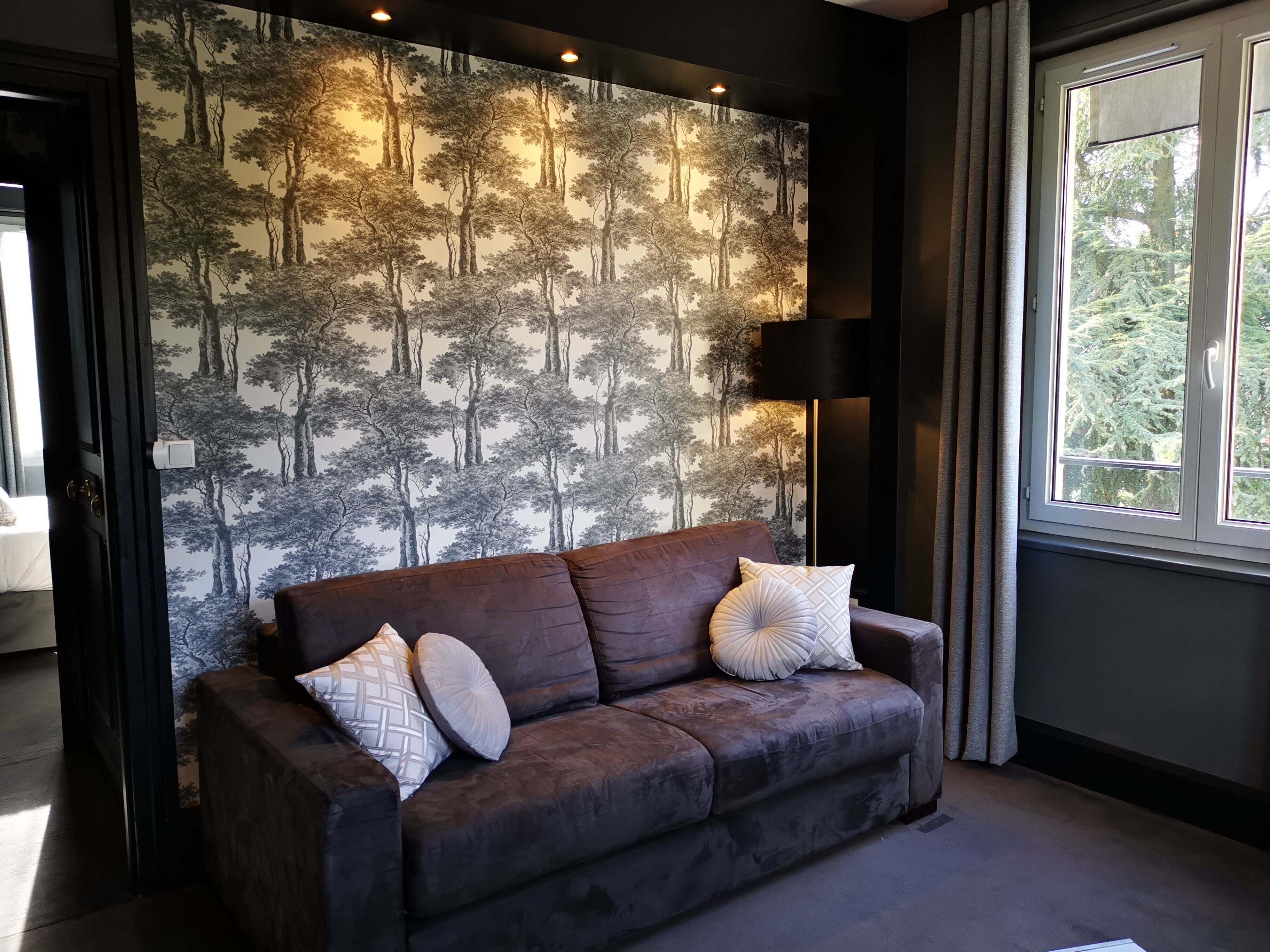 En moderne stue med en mørkebrun sofa, mønstrede puder, væg med skovtapet, gulvlampe og et vindue med gardiner.