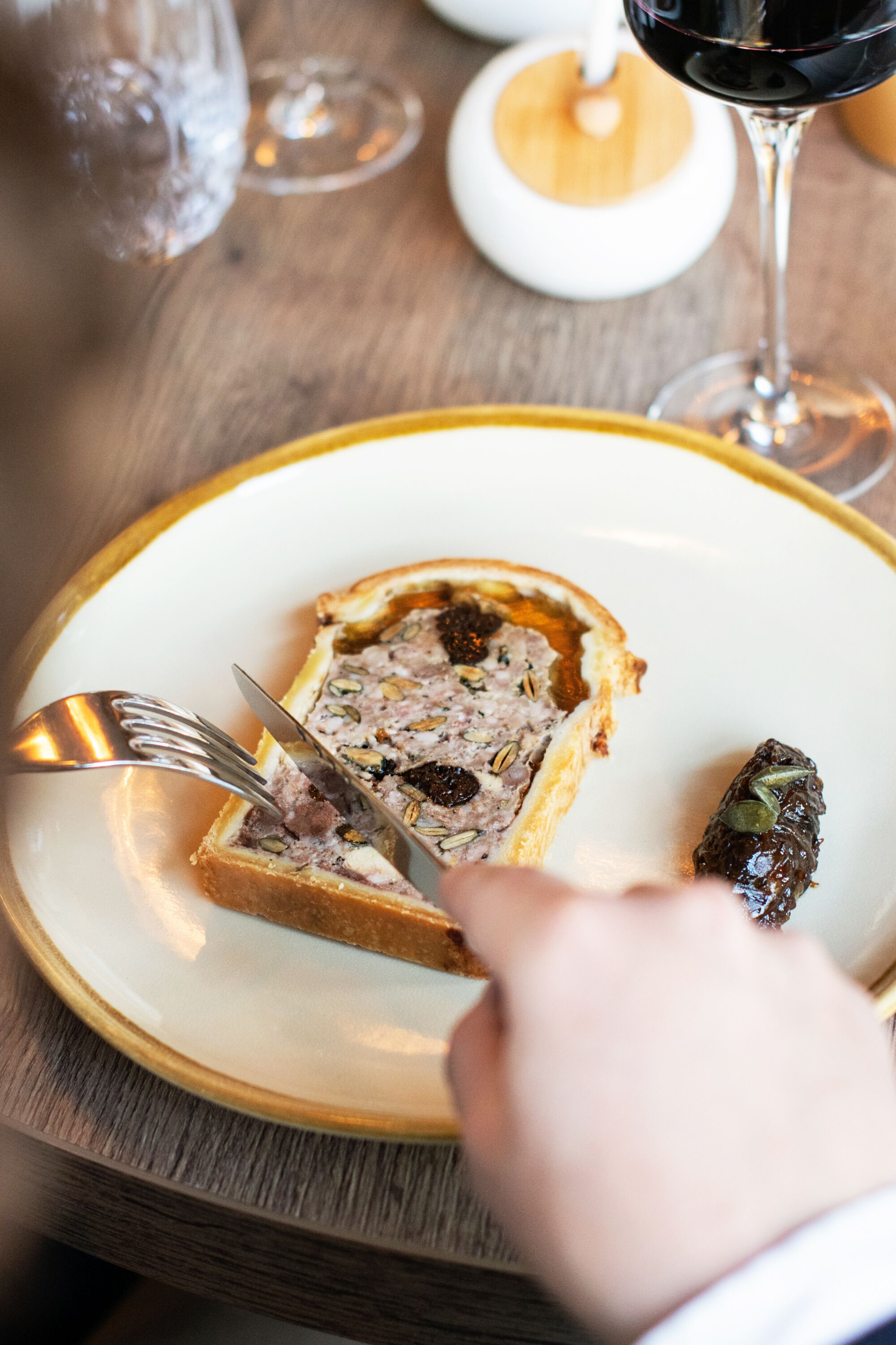En skive kød terrine på en tallerken med en gaffel og kniv, et glas rødvin, og en lille fyldt drue ved siden af.