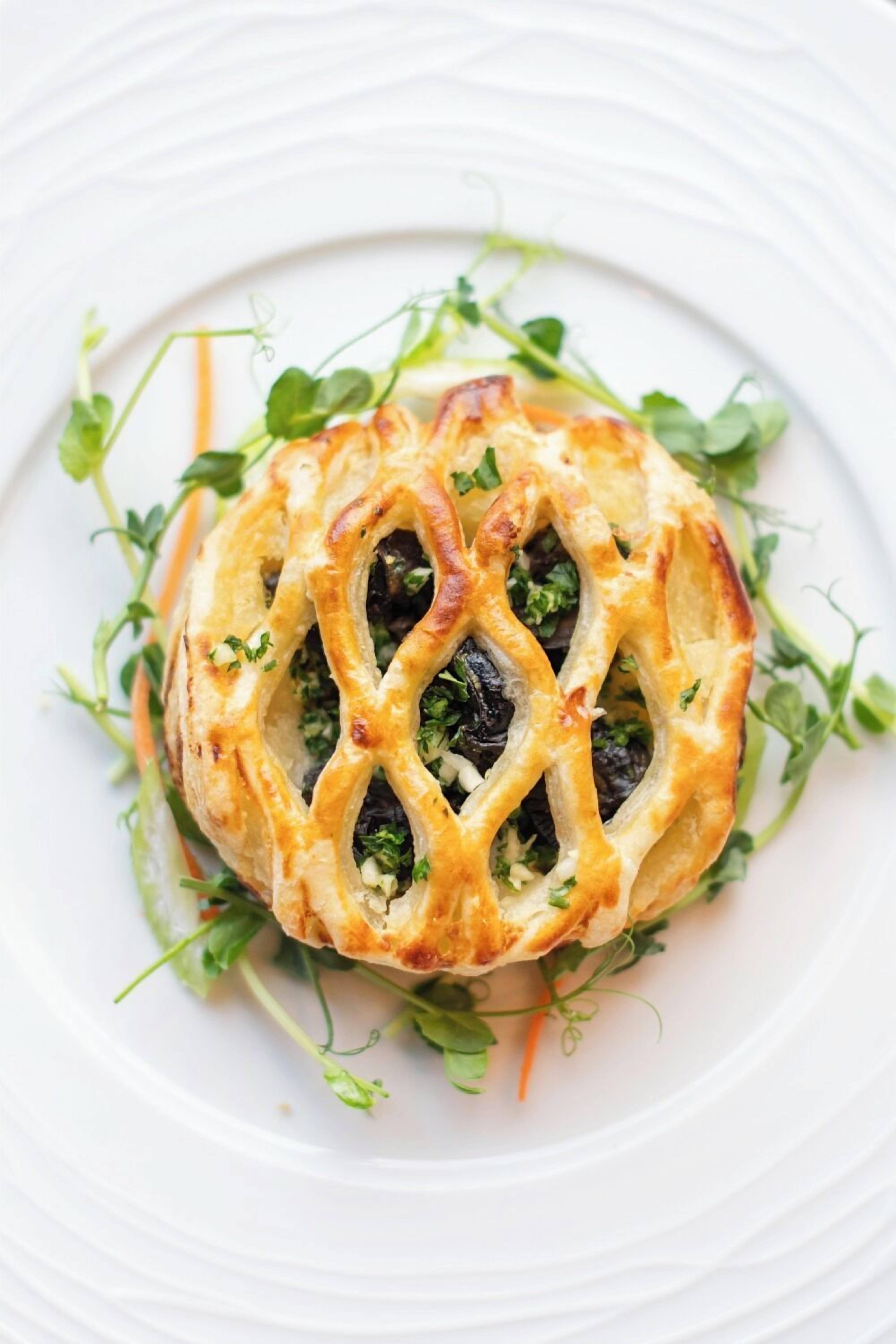 Un plat composé d’une préparation culinaire recouverte d’une pâte feuilletée formant un motif en losanges, décoré de verdure et présenté sur une assiette blanche.