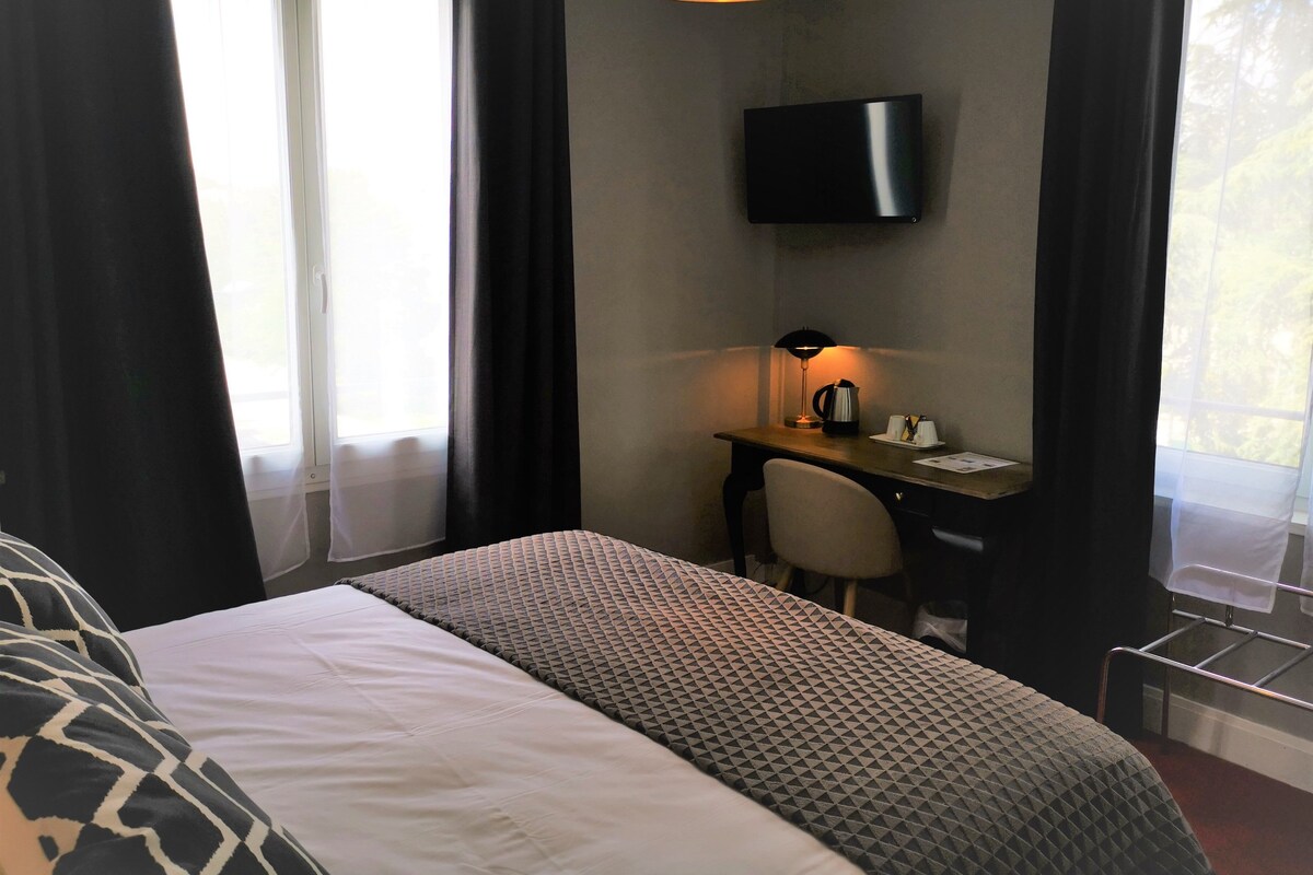 A hotel room with a bed, a desk, a chair, a television mounted on the wall, and a window with curtains.