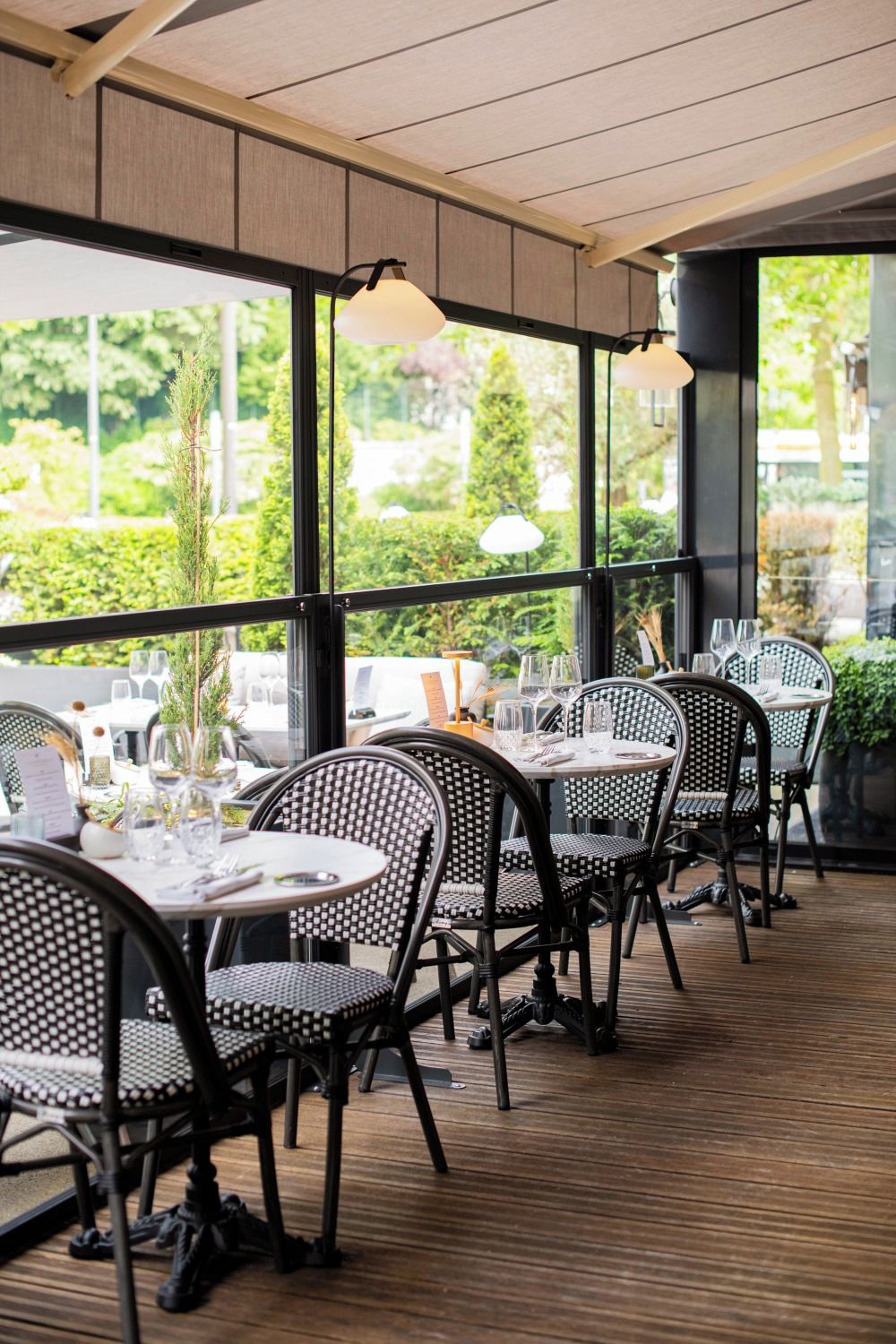 Een eetruimte binnenshuis met tafels en stoelen, grote ramen en uitzicht op groen.