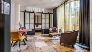 A modern hotel room with a large sliding glass door, a dining area with a wooden table and yellow chairs, a brown leather armchair, and a bed with a black and white striped headboard.