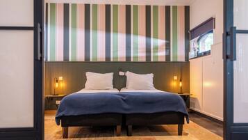 A double bed with a blue bedspread, two white pillows, and a striped wall behind. There are two bedside tables with lamps.