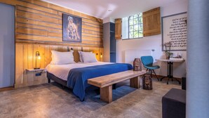 Un dormitorio con cabecera de madera, una cama grande con ropa de cama azul, una mesita de noche con una lámpara, un banco, una cesta de mimbre, una silla y un escritorio con una planta.