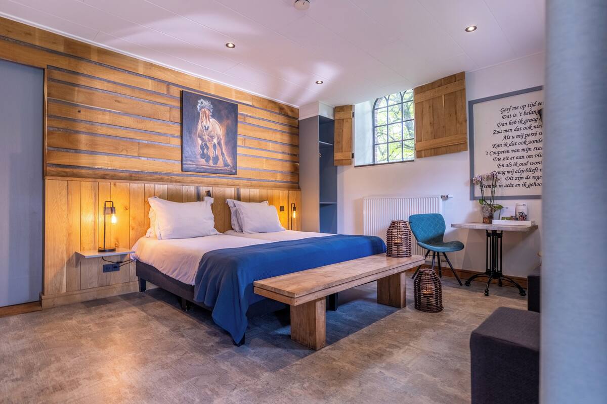 A bedroom with a wooden headboard, a large bed with blue bedding, a bedside table with a lamp, a bench, a wicker basket, a chair, and a desk with a plant.