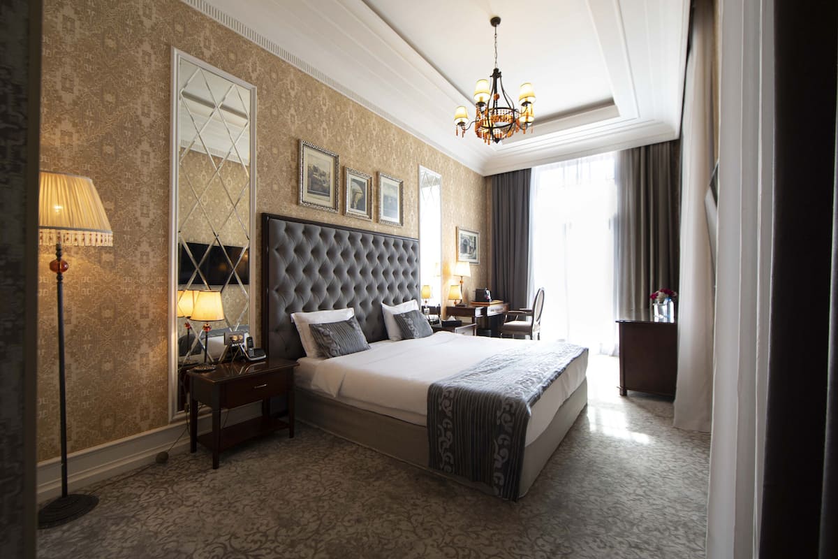 A hotel room with a large bed, a desk, a chair, a lamp, and a chandelier.