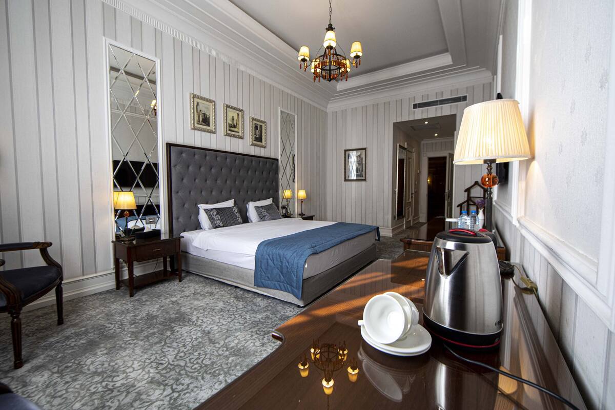 A hotel room with a large bed, bedside lamps, a chandelier, a coffee maker, and a desk with a lamp.