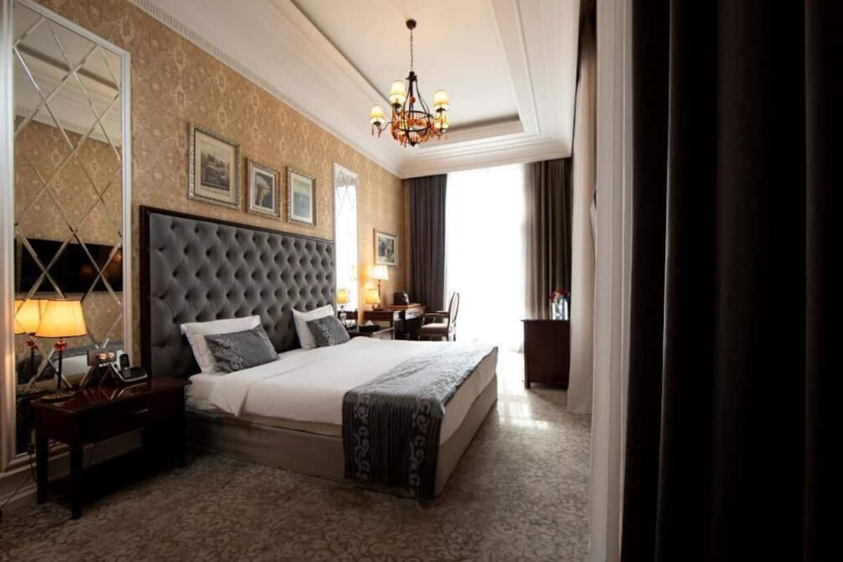 A large bed with a tufted headboard, a bedside table with a lamp, and a mirror. There is a window with curtains, a chandelier, and framed pictures on the wall.