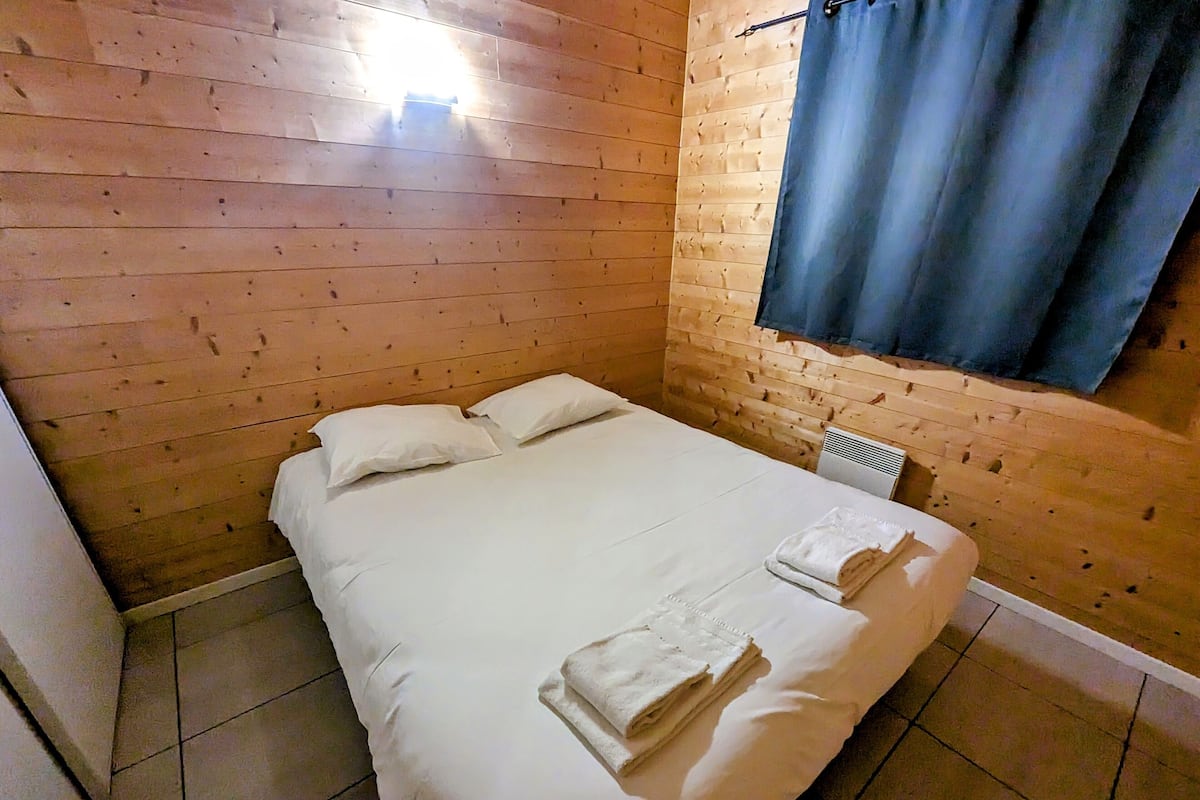 Ein kleines, ordentlich bezogenes Bett mit weißen Leinen und Kissen in einem Raum mit Holzwänden und einem Fenster mit blauen Vorhängen.