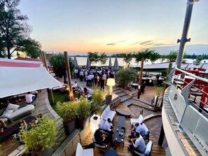 An outdoor dining area with wooden tables and chairs, surrounded by potted plants and umbrellas, overlooking a body of water. - Lodge-Hôtel & Spa les Voiles du Grand Large (Meyzieu)