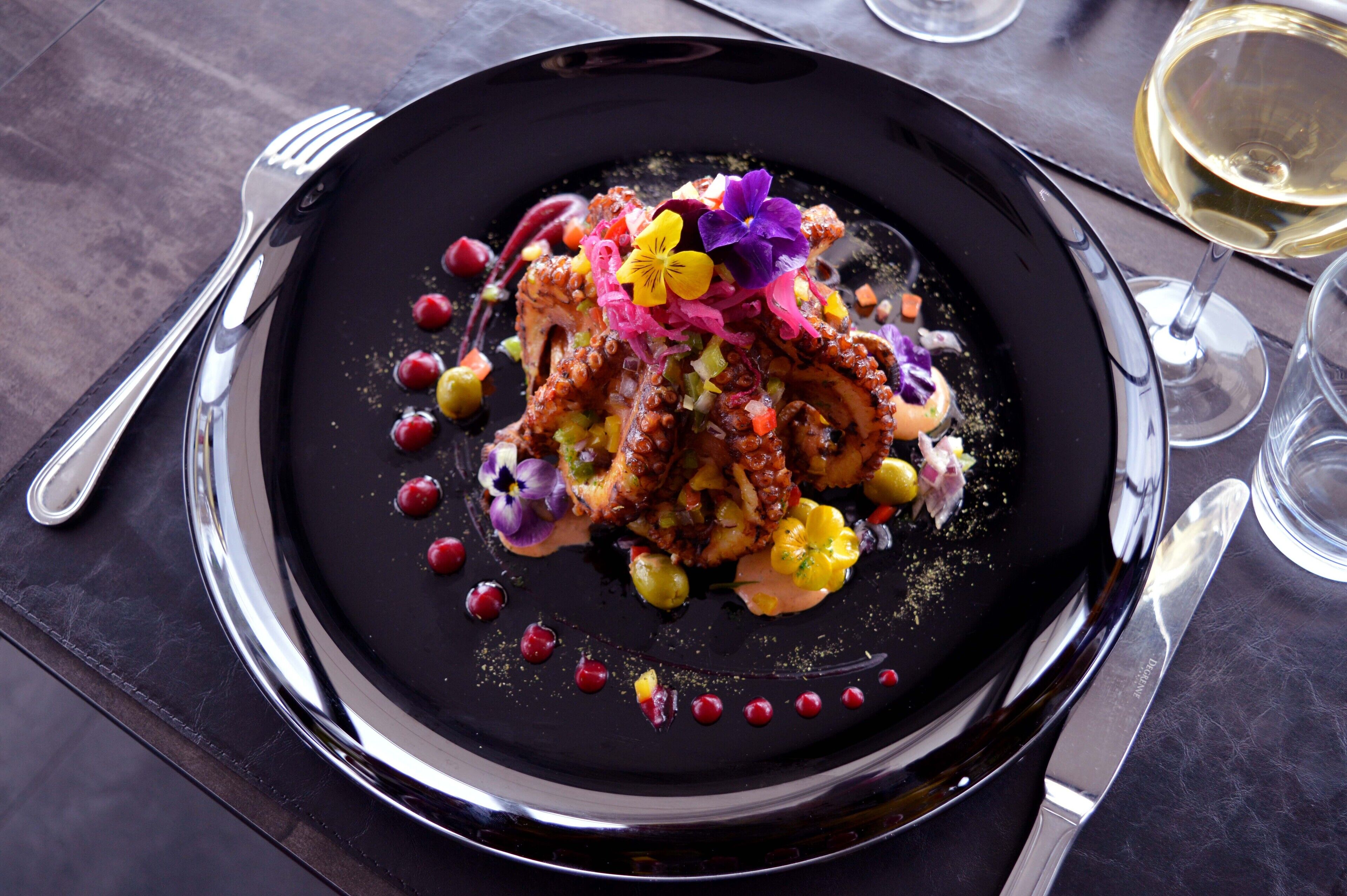 A black plate with a colorful dish, a fork, a knife, and a glass of white wine on a table.