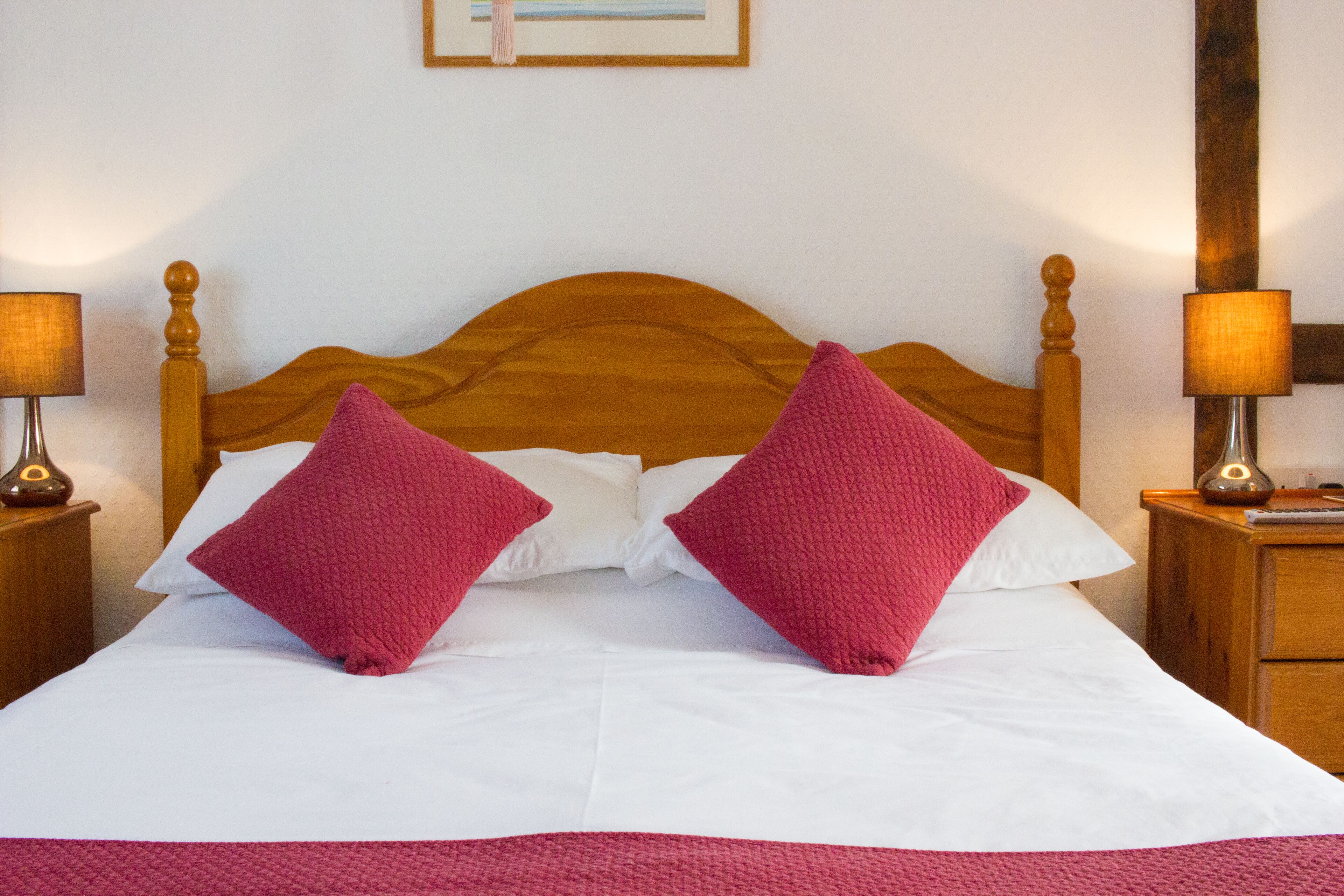 A neatly made bed with white linens and two red pillows, flanked by wooden nightstands with lamps.