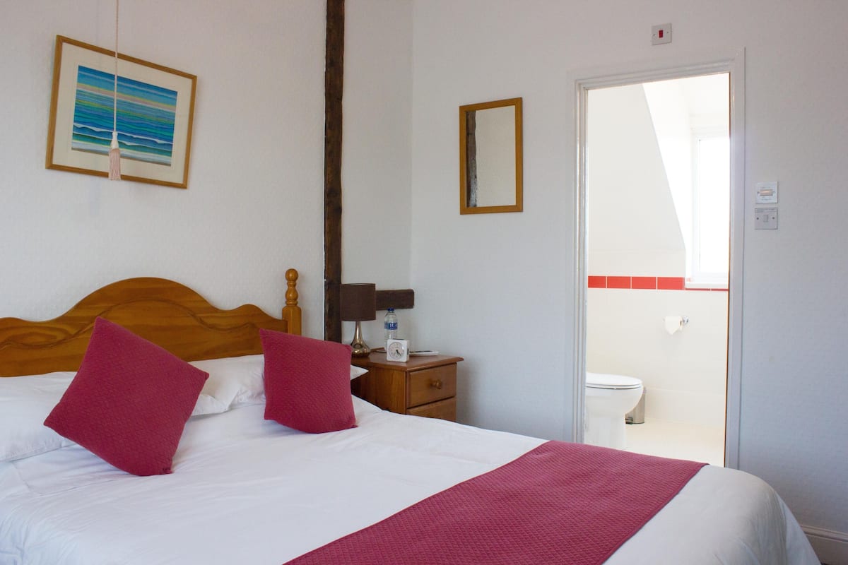 Una cama bien tendida con almohadas rojas, cabecera de madera, mesita de noche con lámpara, un cuadro enmarcado en la pared y un baño visible a través de una puerta abierta.