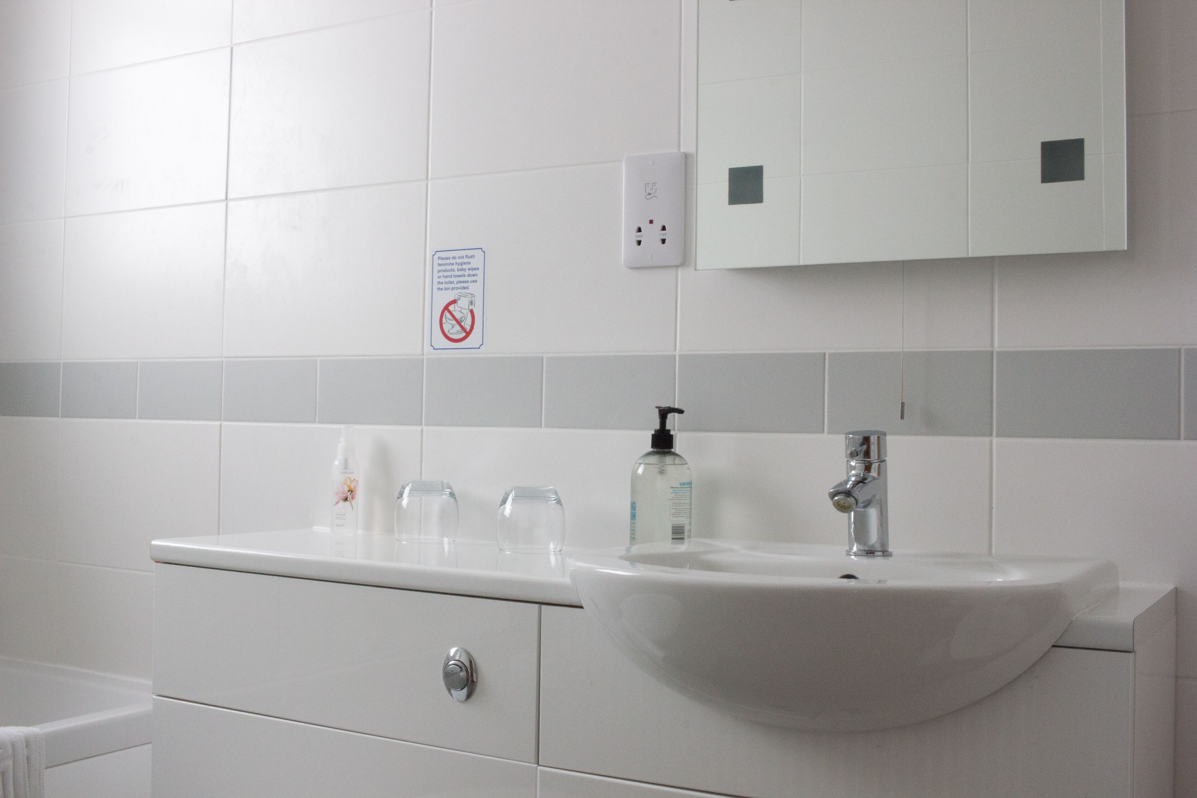 A bathroom with a white sink, mirror, and tiled walls. There are three clear glasses on the sink counter.