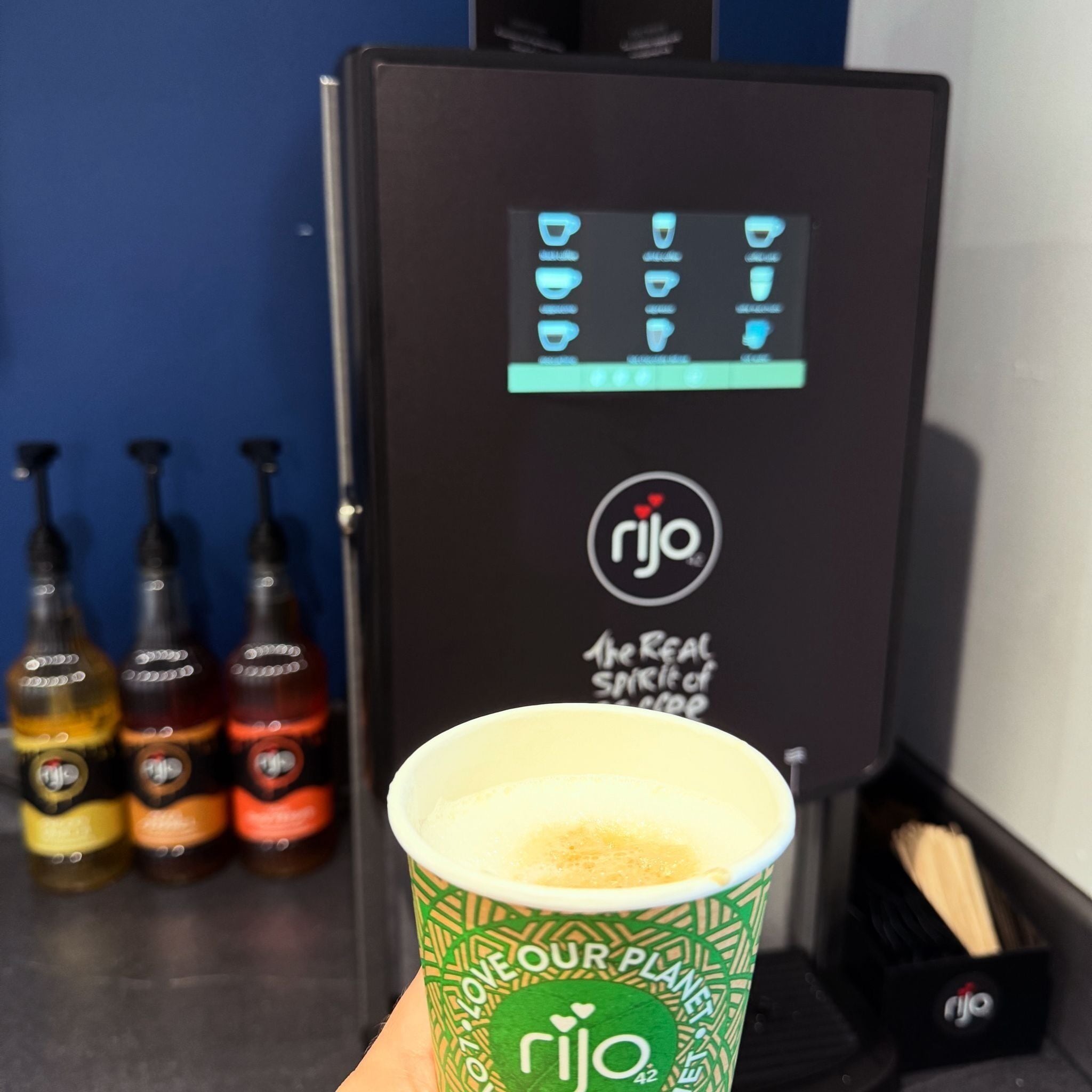 A cup of iced coffee with a green and white design, a beverage dispenser with the logo 'Rijo,' and three bottles of different colored drinks in the background.