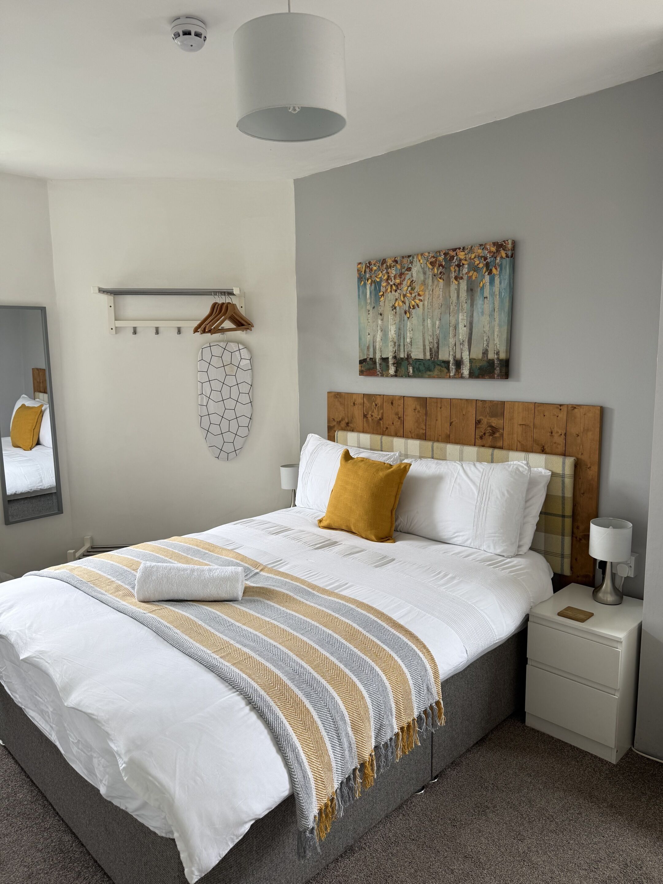 Ein ordentlich eingerichtetes Schlafzimmer mit Bett, Nachttischen, einem Kleiderhaken, einem Wandbild und einer Deckenleuchte.