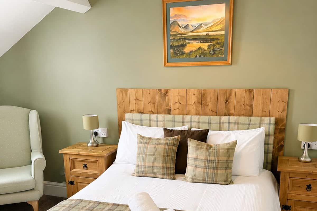 A neatly made bed with a plaid bedspread and pillows, a wooden headboard, a painting on the wall, and a chair in a room with green walls.