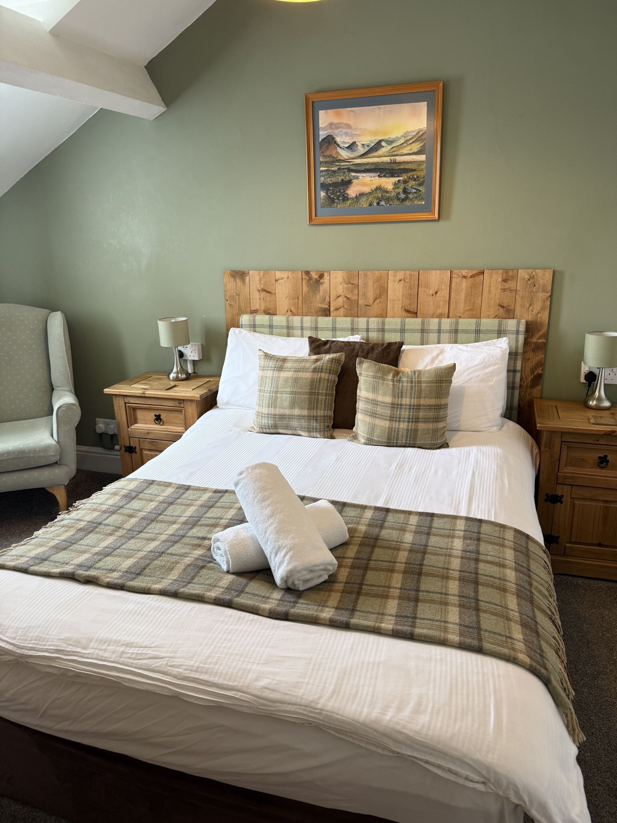 A neatly made bed with a plaid blanket and pillows, a wooden headboard, a framed landscape painting, and a chair in a room with green walls.