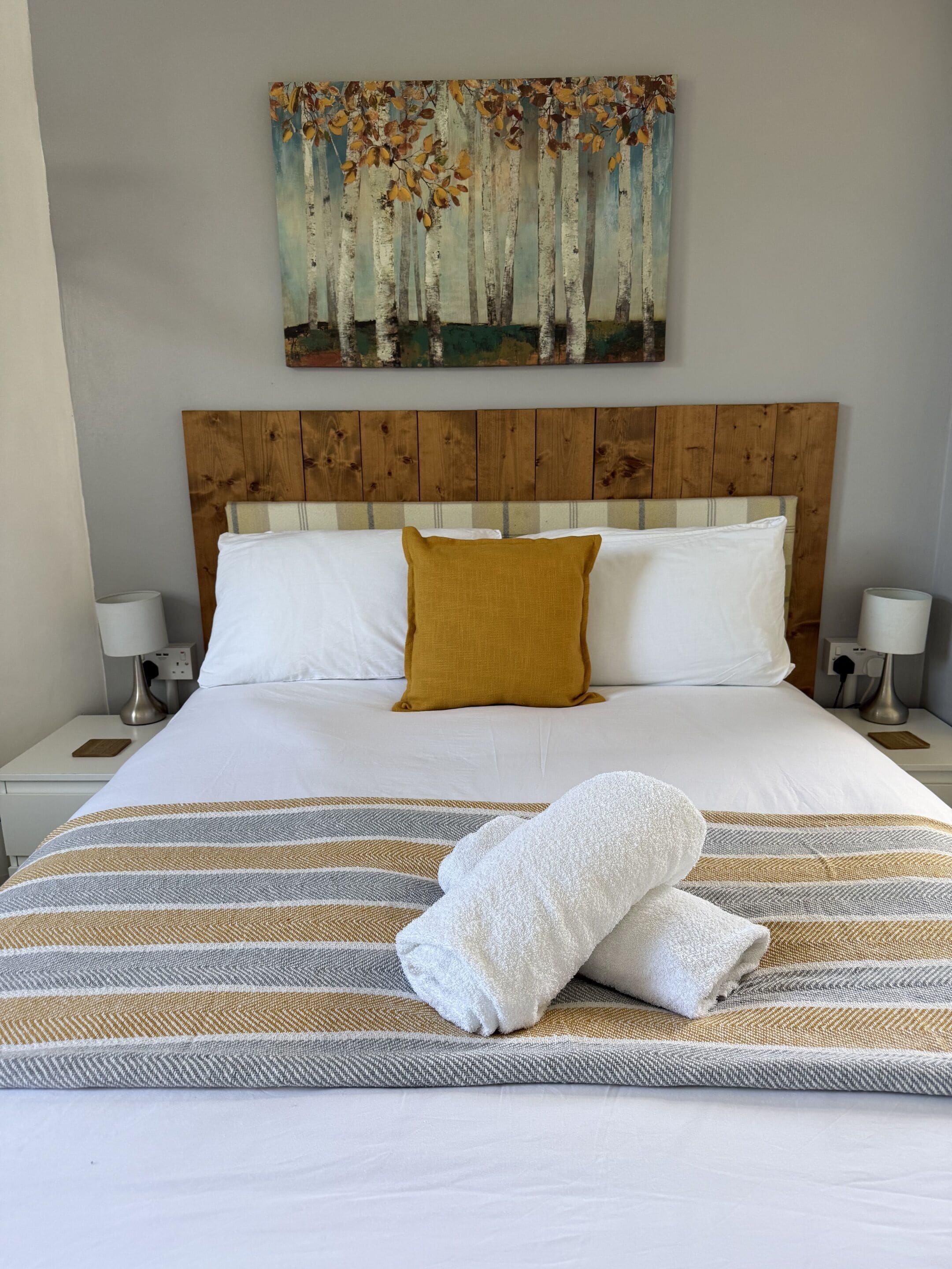A neatly made bed with white and mustard pillows, a striped area rug, and a painting of birch trees on the wall.