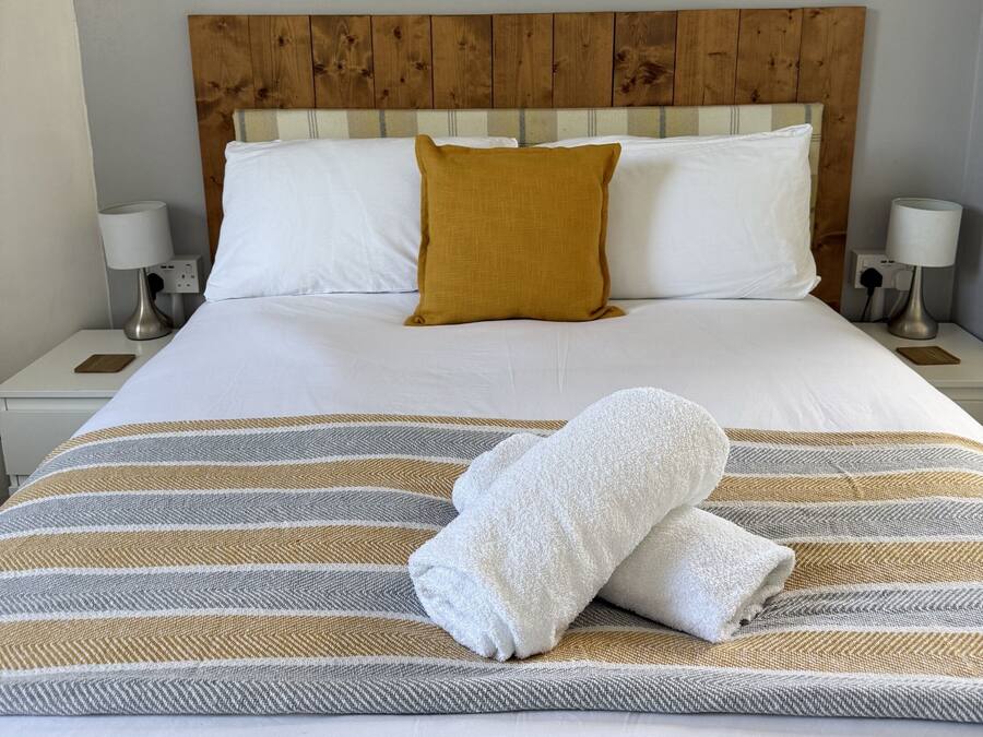 A neatly made bed with white and mustard pillows, a striped area rug, and a painting of birch trees on the wall.
