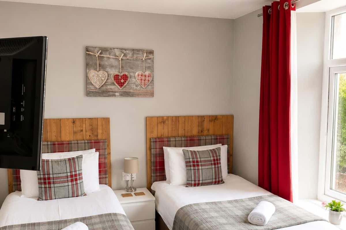 A hotel room with two beds, a TV, a desk with a chair, a lamp, and a window with red curtains.