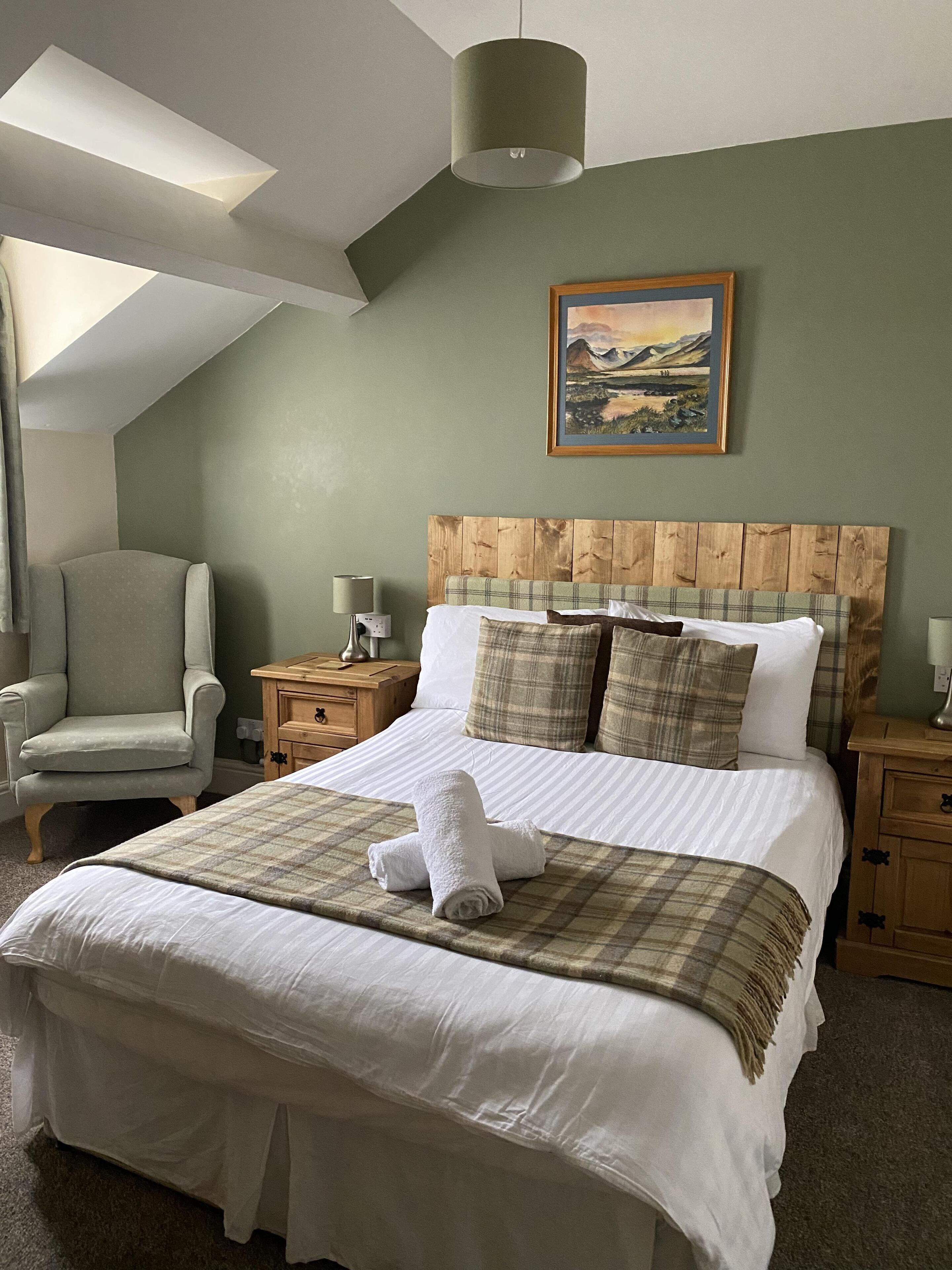 A neatly made bed with a plaid blanket and rolled towels, a wooden headboard, a painting on the wall, a chair, and a bedside table with a lamp.