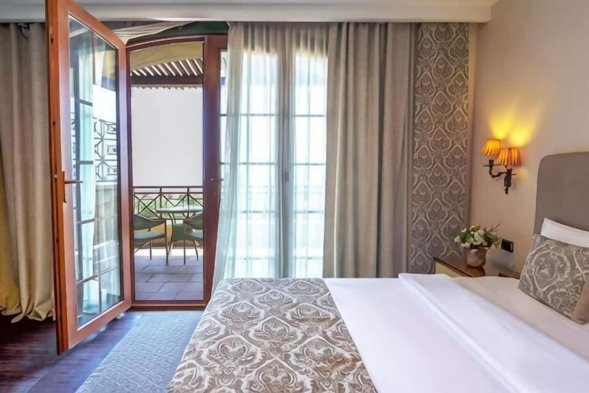 A hotel room with a bed, a nightstand with a lamp, a view of a balcony with a table and chairs, and a patterned wall.