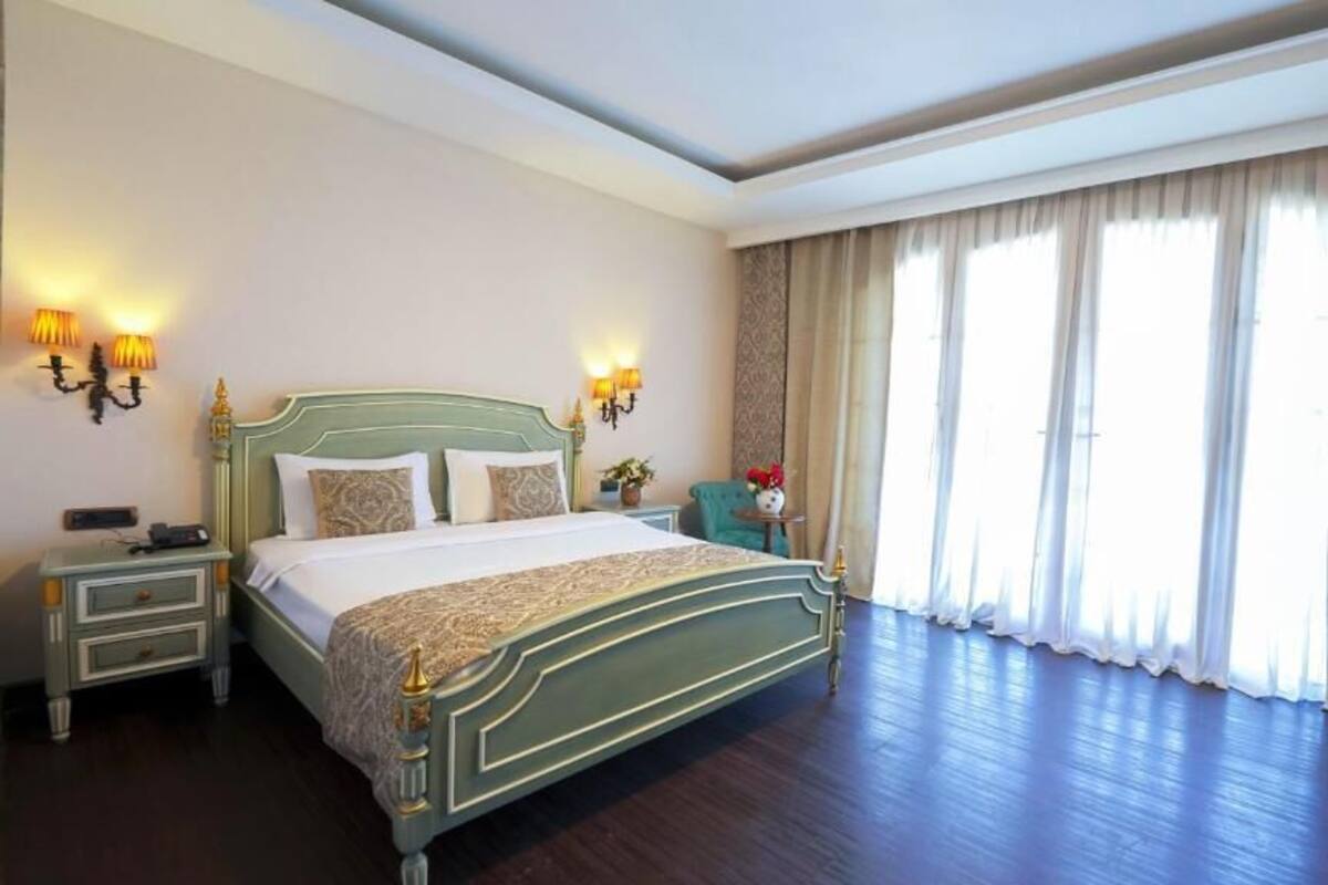 A spacious hotel room with a large bed, two bedside tables, a chair, and a large window with curtains.