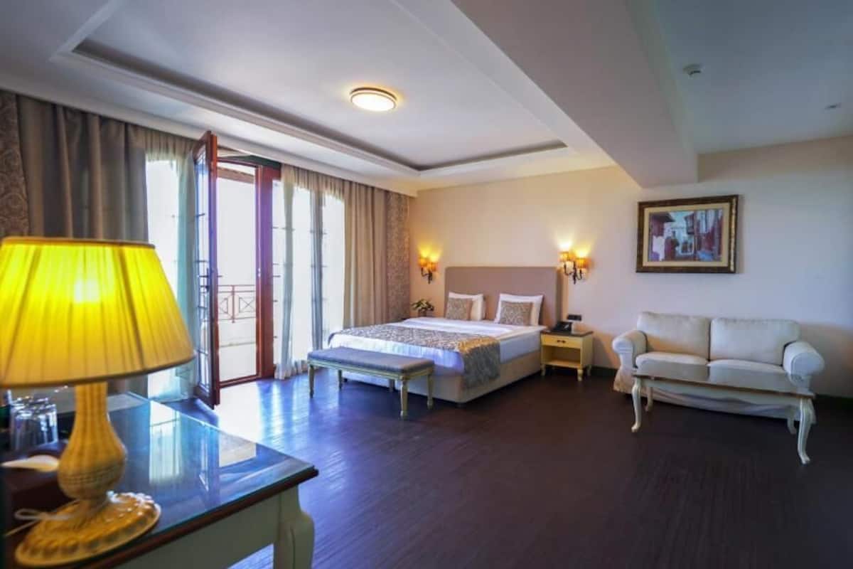 A hotel room with a bed, bedside tables, a sofa, a desk with a lamp, a wardrobe, and a painting on the wall.